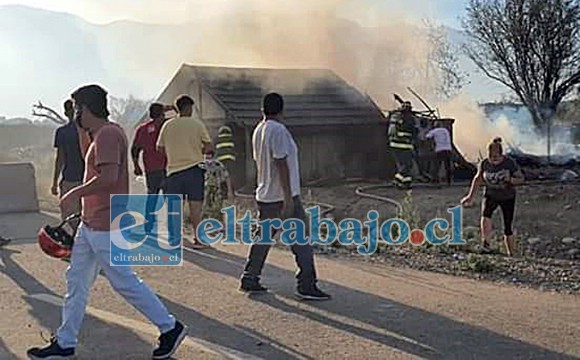 Bomberos trabajando en el lugar, mientras vecinos observan la vivienda siniestrada.